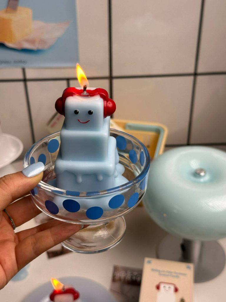 Whimsical pastel blue melting ice cube character soy candle wearing red headphones displayed in dessert glass cup.